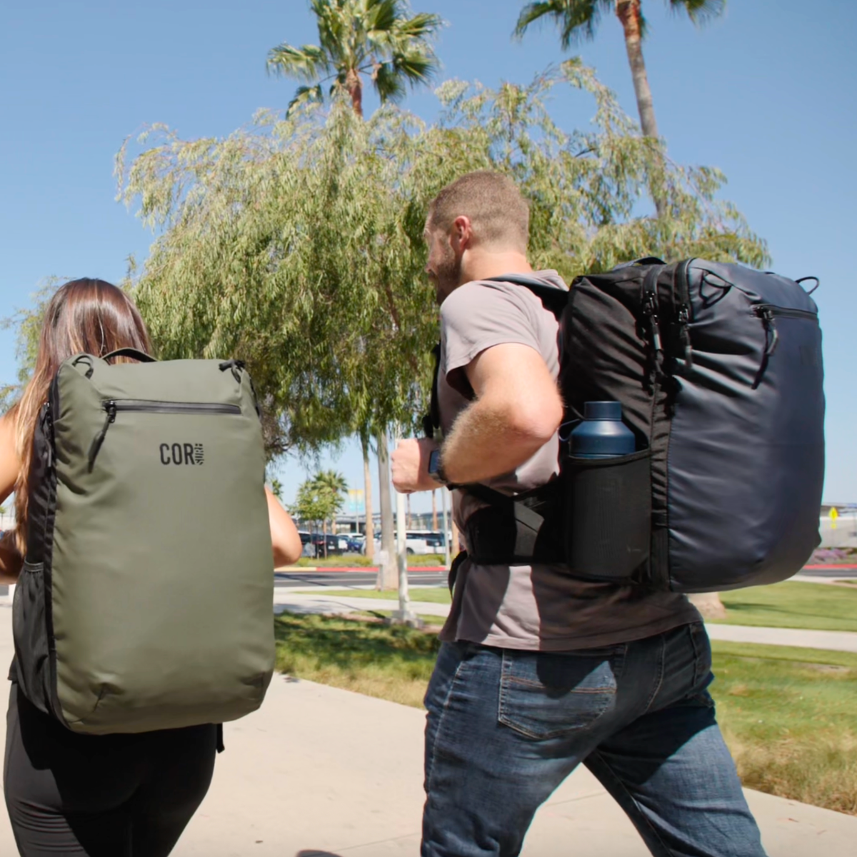 Two travelers carrying green and blue Island Hopper backpacks outdoors, showing the bags from the back and side.