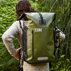 Person wearing a green COR Surf dry bag backpack among trees, with a bottle in the side pocket.