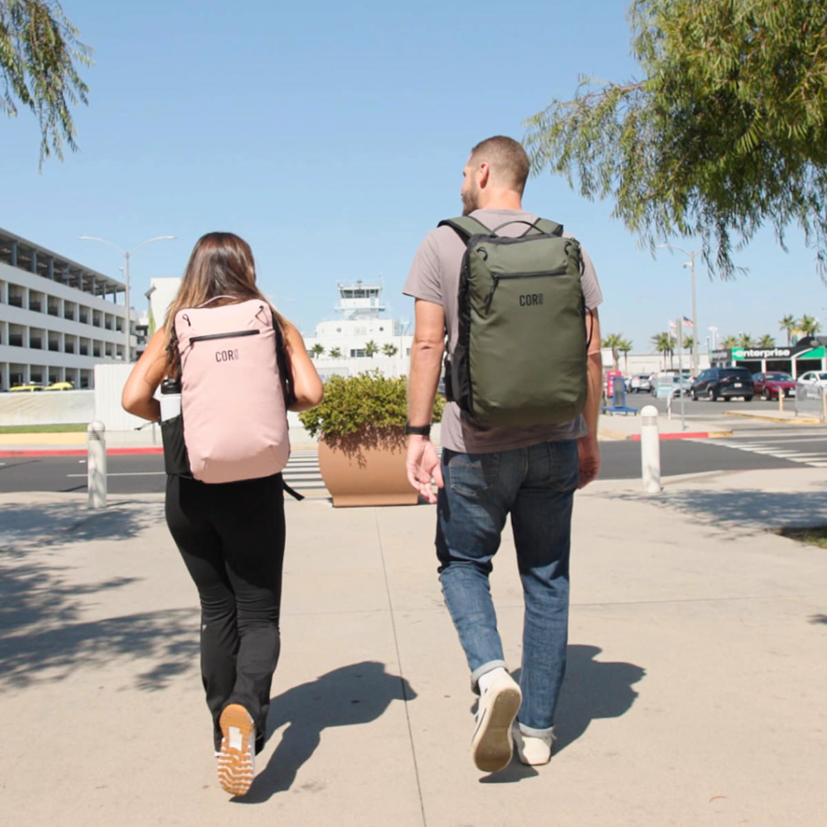 Two travelers wearing pink and green COR Surf backpacks outside an airport.